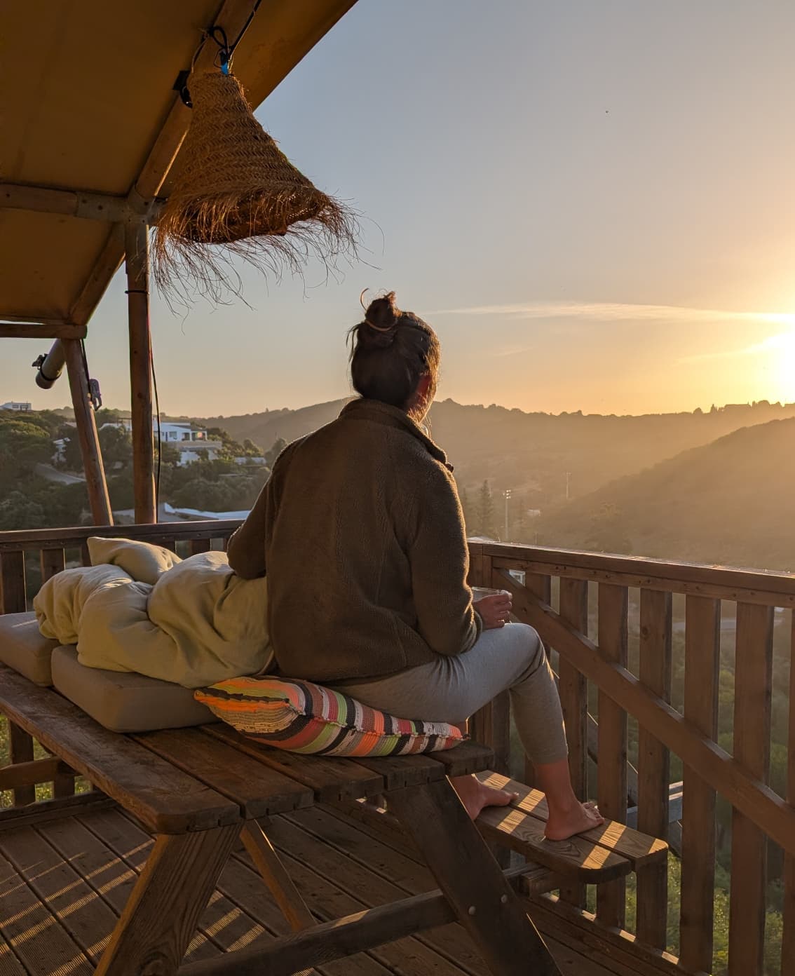 Frau sitzt barfuß auf einer Holzbank auf einer Terrasse und blickt über bewaldete Hügel in den goldenen Sonnenuntergang Portugals.