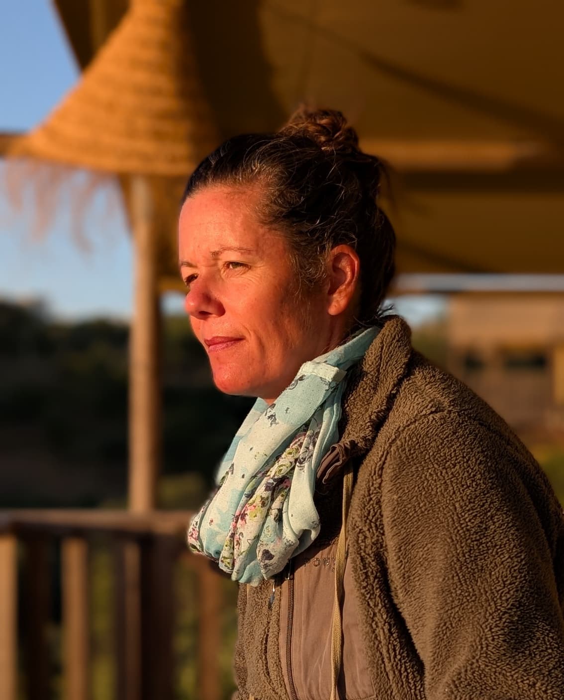 Dr. Veronika Klein im Profil bei Sonnenuntergang, nachdenklich in die Ferne blickend, mit Schal und Fleecejacke auf einer Terrasse in Portugal.