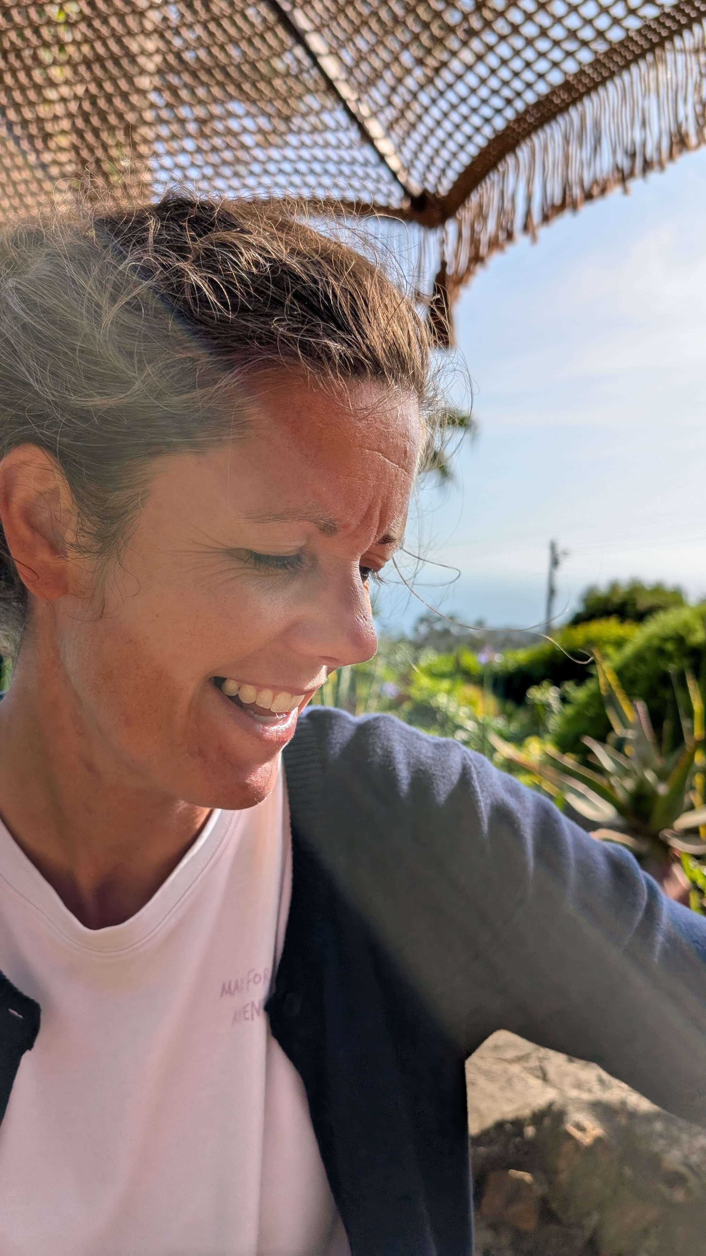 Dr. Veronika Klein lächelt entspannt im Sonnenlicht unter einem Sonnenschirm, im Hintergrund ein grüner Garten.