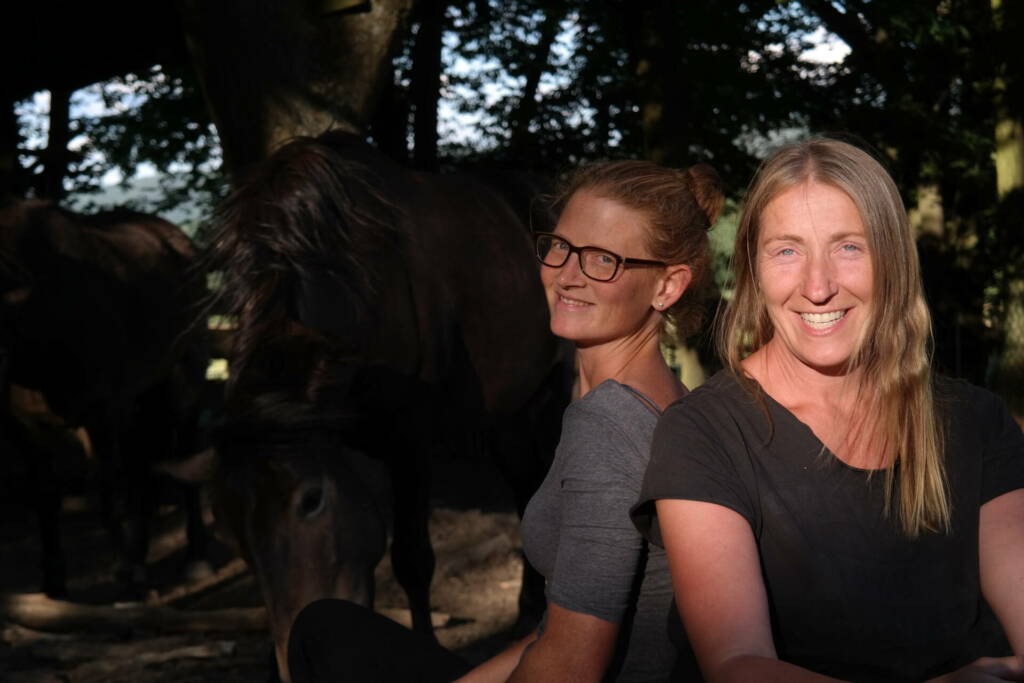 Christina und Barbara von Team HUF sitzen Rücken an Rücken, im Hintergrund grasen dunkle Pferde im Abendlicht