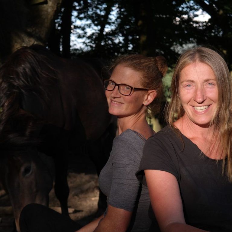 Christina und Barbara von Team Huf sitzen lächelnd vor einem dunklen Pferd im Schatten von Bäumen