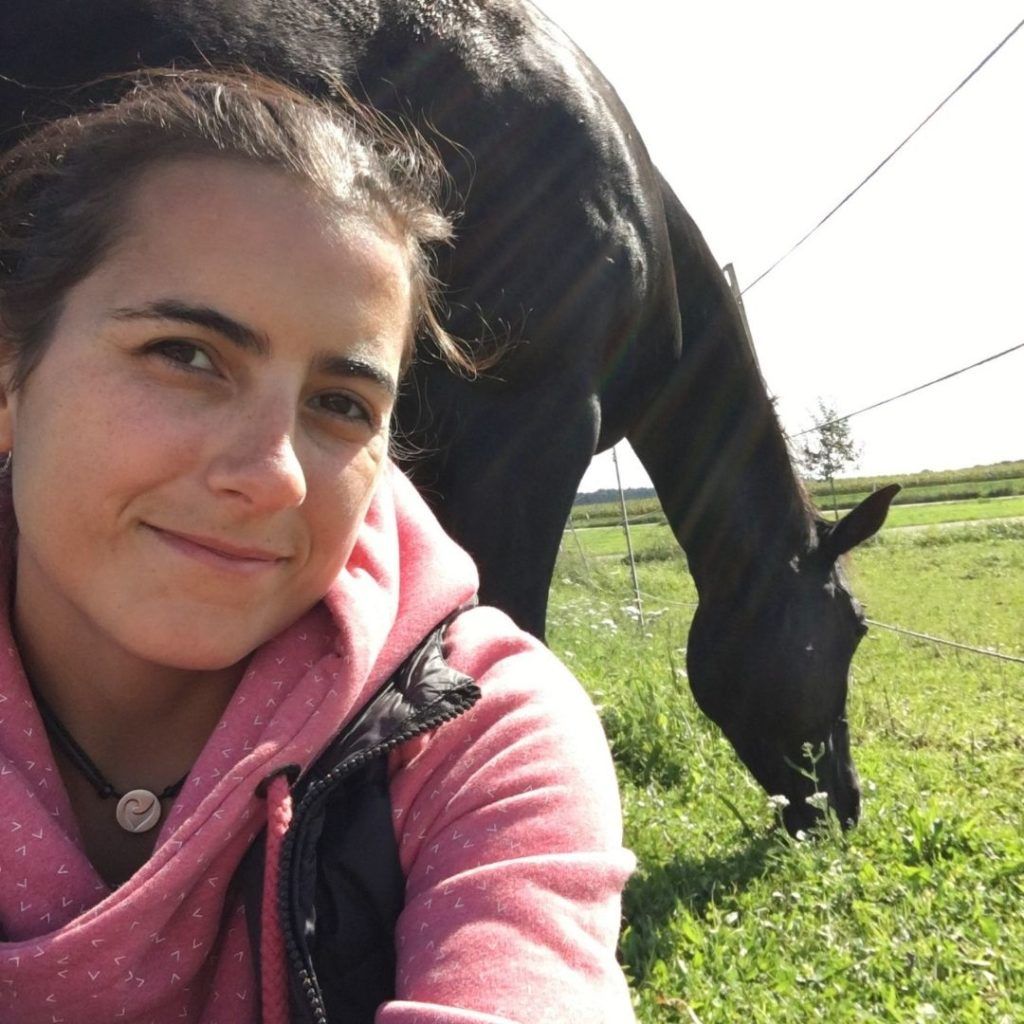 Junge Frau macht ein Selfie auf der Weide, im Hintergrund grast ihr schwarzes Pferd
