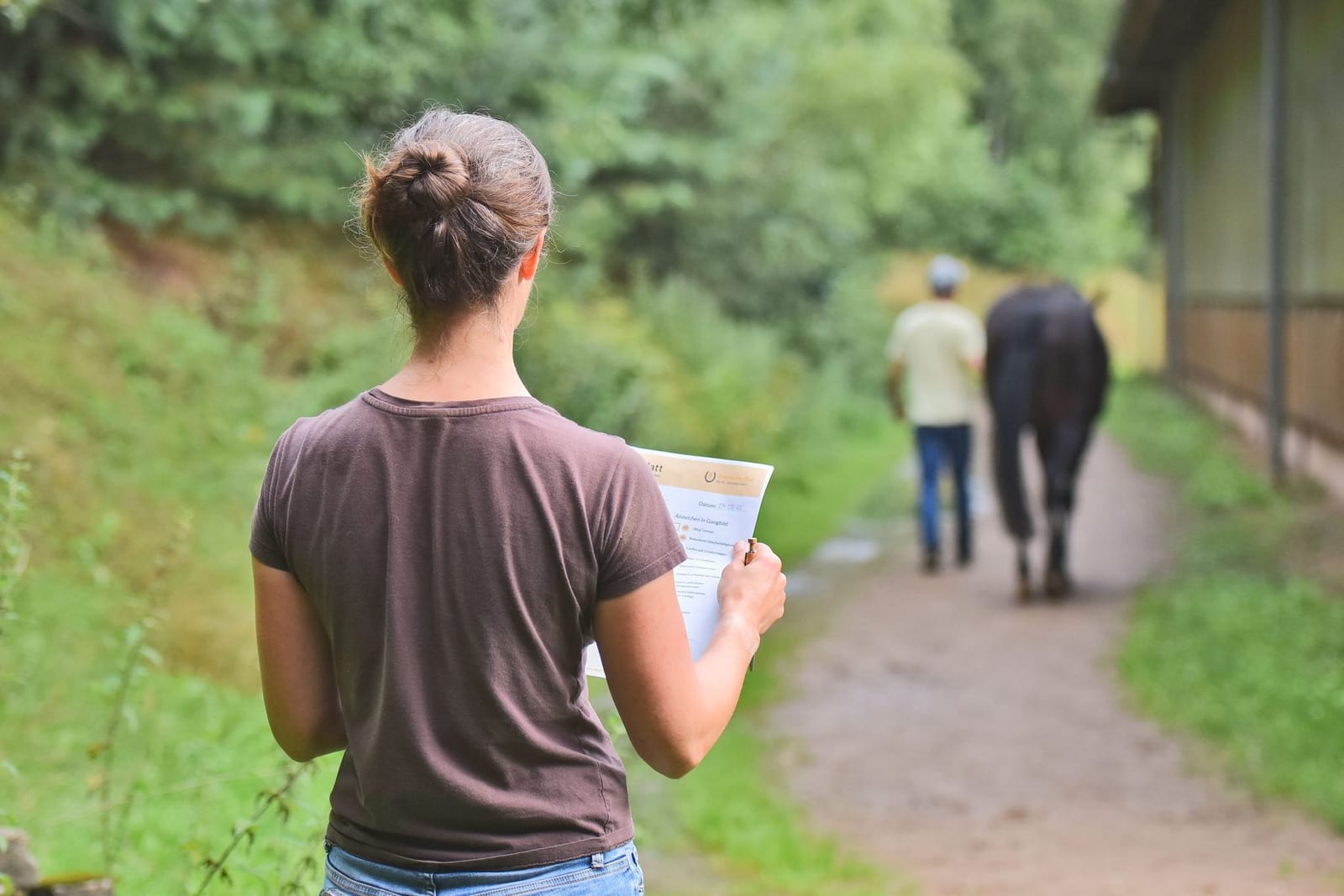 Pferdebesitzerin mit Beobachtungsbogen schaut einem Pferd nach, das auf einem Feldweg von ihr weggeführt wird